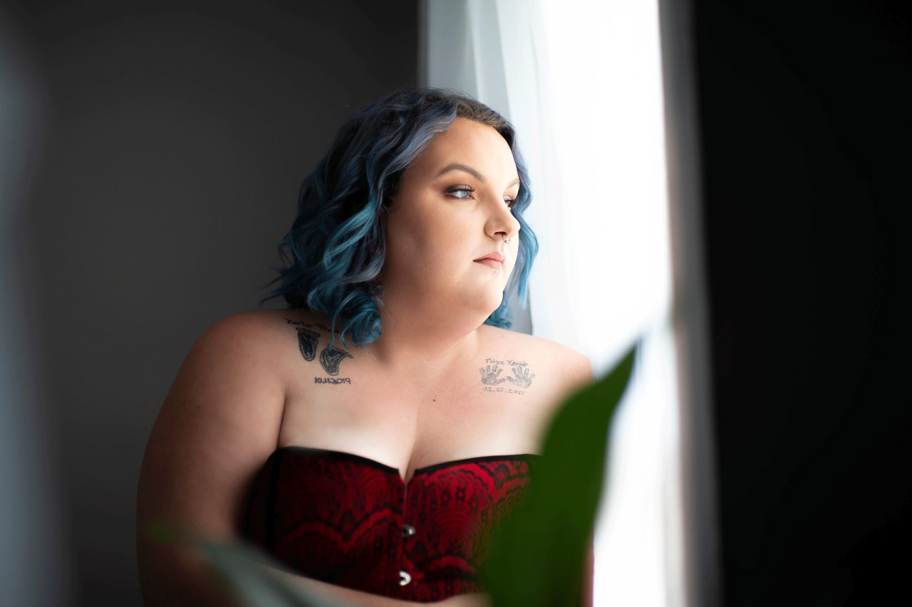 Blue haired woman wearing a red corset looks out a window for her boudoir photoshoot