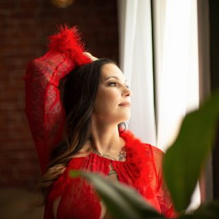 Young female wearing a red robe looks out a window during her plus size boudoir photography session