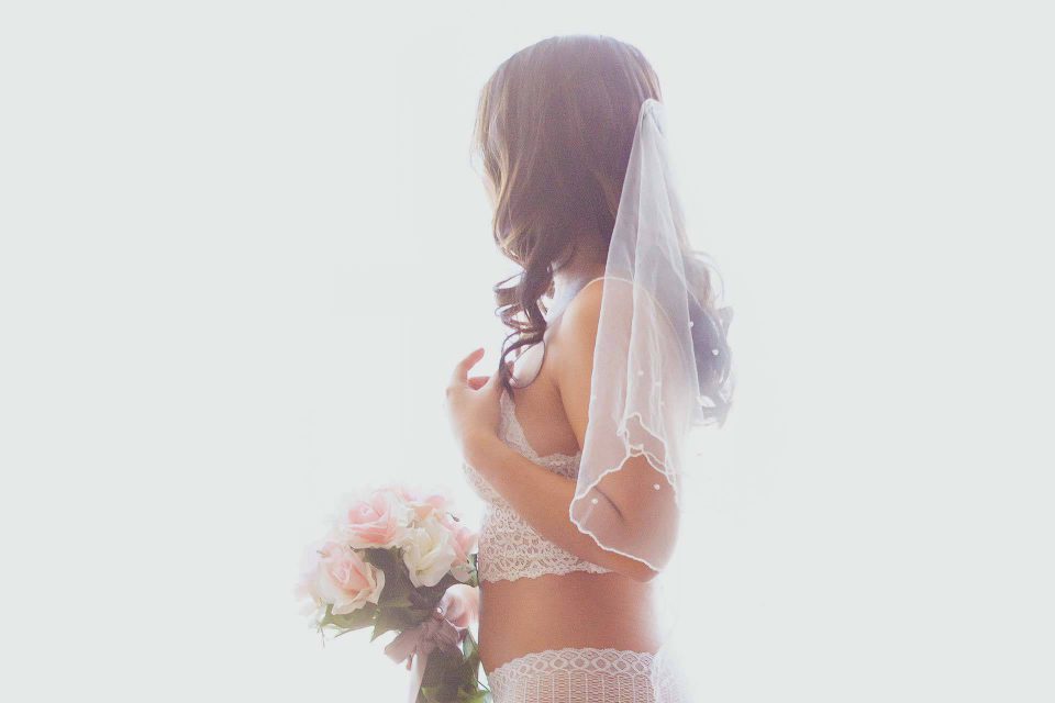 dark haired woman gently holding light pink and white rose bouquet in white lace set and veil poses for wedding boudoir photography in Tampa, FL